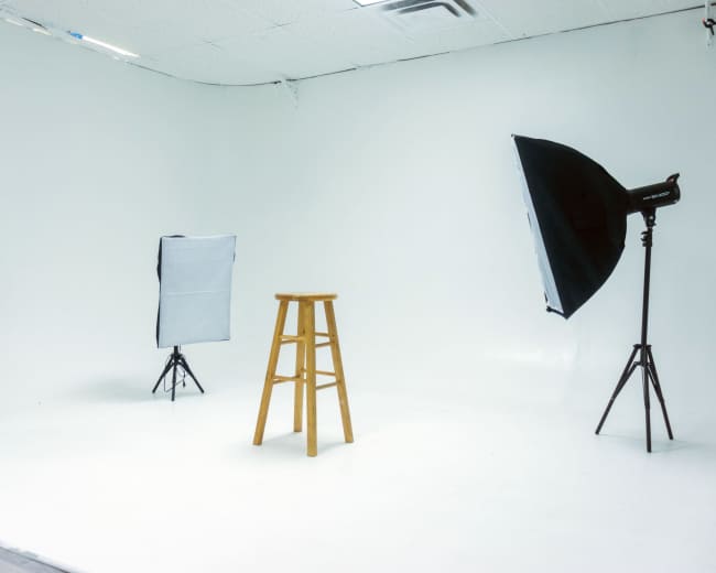 A photography studio features a wooden stool positioned in the center, surrounded by three softbox lights on tripods.