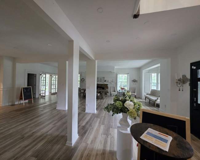 The image shows a spacious, well-lit interior with open areas, wooden flooring, and several furnishings including a floral arrangement and seating.