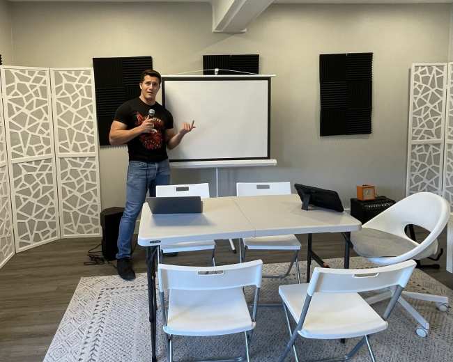A person stands beside a projection screen in a modern meeting room with a table and chairs arranged for a presentation.