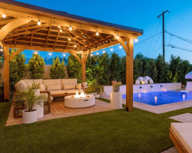 The image shows a landscaped outdoor area featuring a wooden gazebo with seating, a fire pit, a swimming pool, and lounge chairs under a twilight sky.