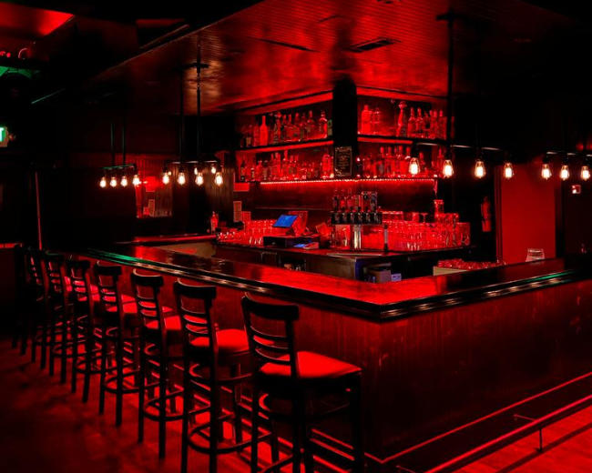 A dimly lit bar features a red and black color scheme, with a long counter and several hanging light fixtures above.