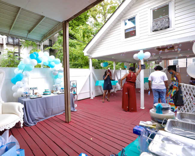 A backyard party setup features a table with decorations and treats, balloons, and guests interacting under a covered patio.