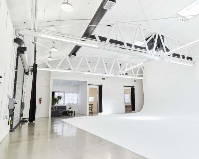 The image shows a spacious, bright studio with white walls, a seamless backdrop, and large windows allowing natural light.