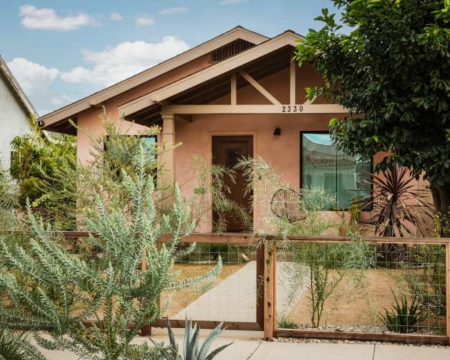 The image shows a single-story house with a light brown exterior and a wooden fence surrounding a garden with various plants.