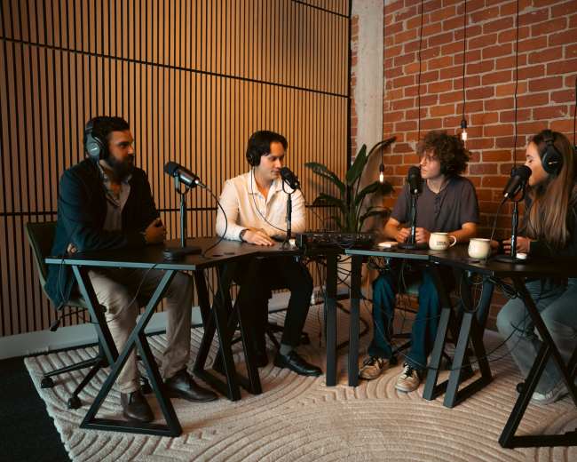Four people sit at a table in a well-lit room with exposed brick walls, engaged in a podcast discussion, each using a microphone.