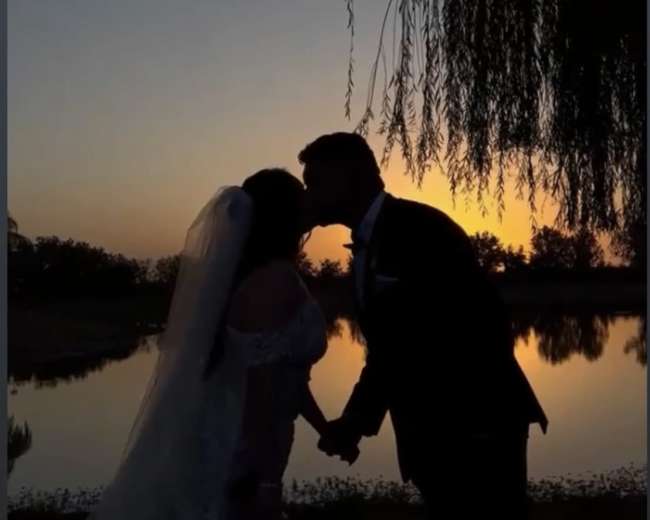 A couple shares a kiss at sunset near a water body, framed by a willow tree.