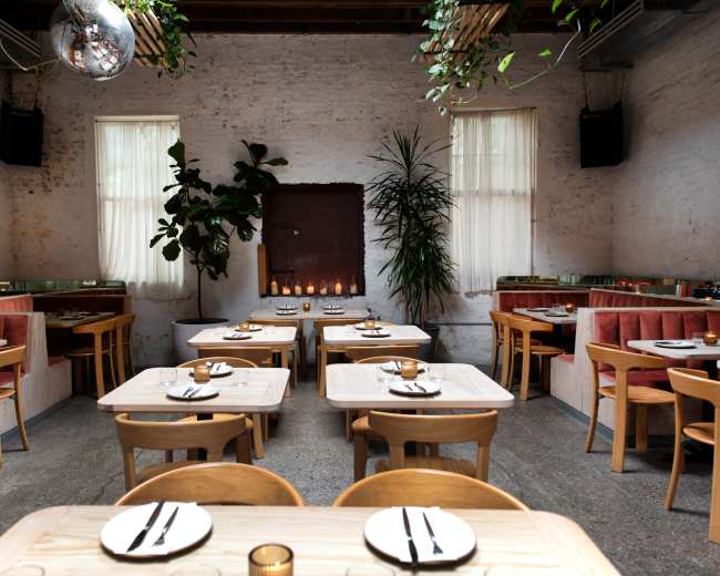 A modern restaurant interior features wooden tables and chairs, with plants and candles adding decor along the walls.