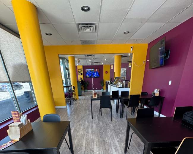The image shows a modern café interior featuring black tables and chairs, colorful purple and yellow walls, and a large television on the wall.