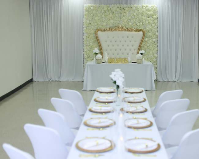 A long dining table with white chairs is set up in a elegantly decorated room featuring a floral backdrop and a gold-accented head table.