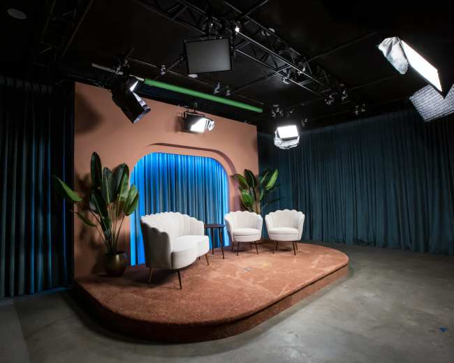 The image shows a modern set with three white chairs on a brown carpet, flanked by tall green plants and illuminated by multiple studio lights against a backdrop of blue and pink curtains.