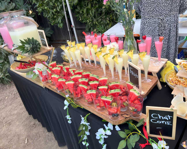 A table display featuring a variety of colorful drinks in clear glasses, garnished with fruit, along with two large beverage dispensers and snacks.