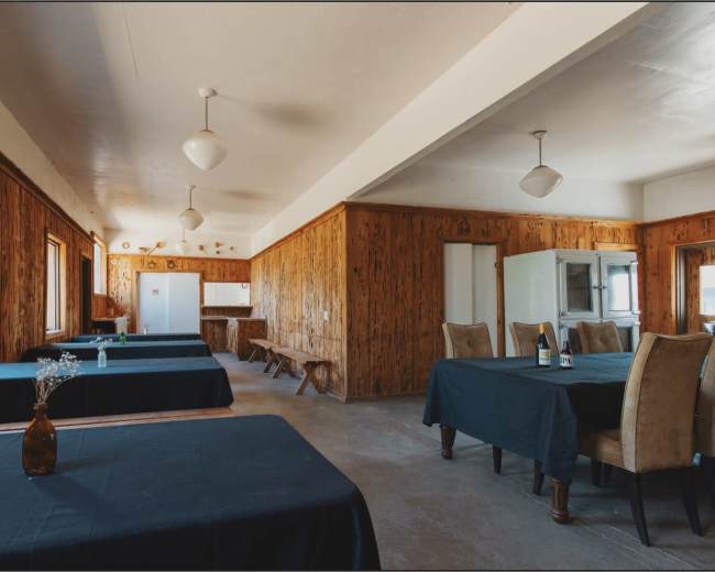 A spacious, sparsely furnished dining area with wooden walls, several tables covered in dark tablecloths, and simple light fixtures hanging from the ceiling.