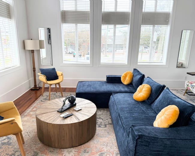 The living room features a dark blue sectional sofa with yellow pillows, an oval wooden coffee table, and large windows that provide ample natural light.