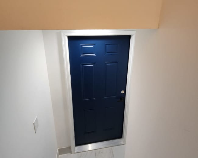 A blue door with six panels is located at the bottom of a staircase, framed by white walls and a ceiling.