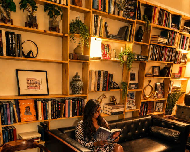 A woman sits on a black leather couch in a cozy library, reading a book surrounded by wooden shelves filled with books and plants.