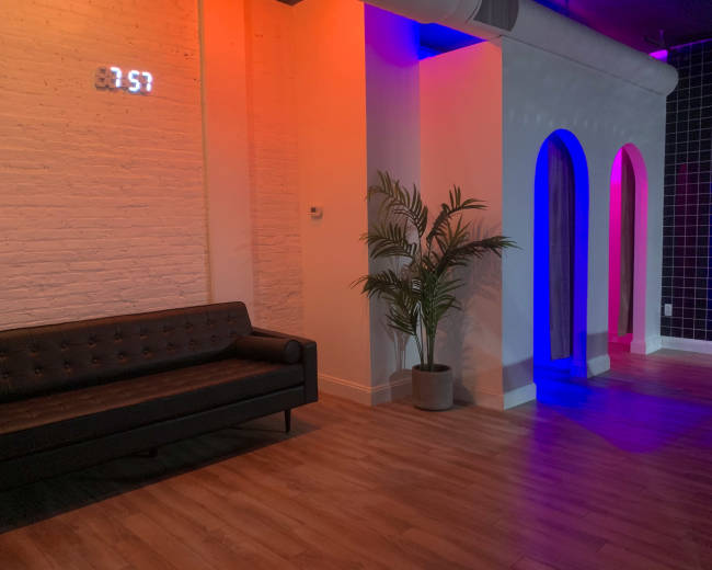 The image shows a modern interior space featuring a black couch, a potted plant, and colorful LED lighting along the walls and archways.
