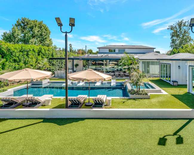 The image shows a modern home with a spacious outdoor area featuring a swimming pool, lounge chairs, and umbrellas, surrounded by greenery and well-maintained landscaping.