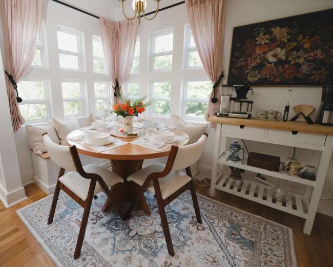 A round table is set for a meal in a brightly lit dining area with large windows and floral arrangements.