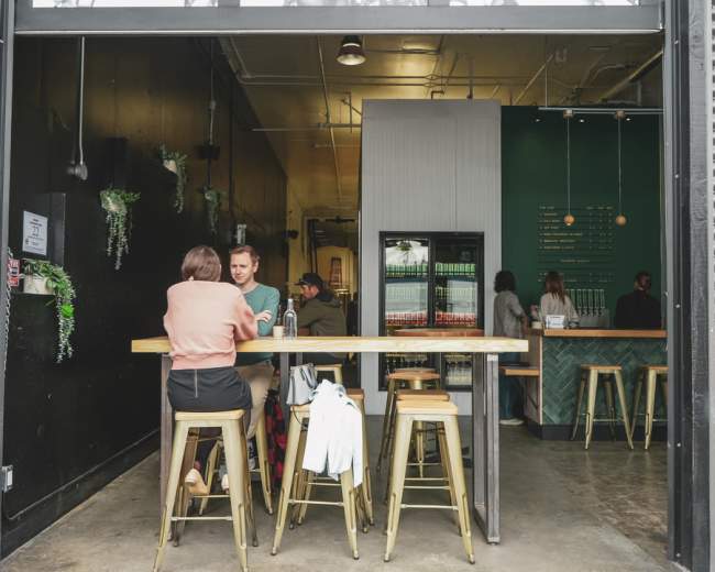 Two people sit at a long wooden table inside a modern café, with a green wall and a beverage fridge visible in the background.