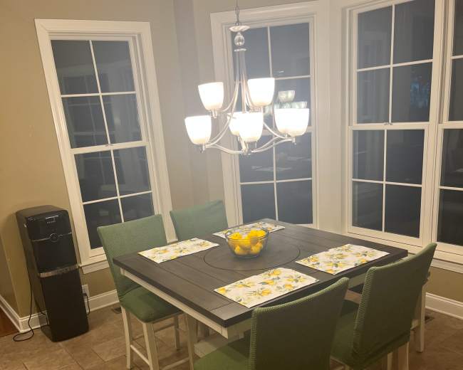 A wooden dining table with six green chairs is set with placemats and a bowl of lemons, surrounded by large windows.
