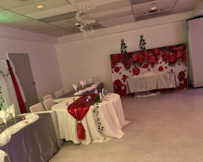 A decorated event space featuring tables covered with white and silver tablecloths, accented by red decorations and floral motifs on the walls.