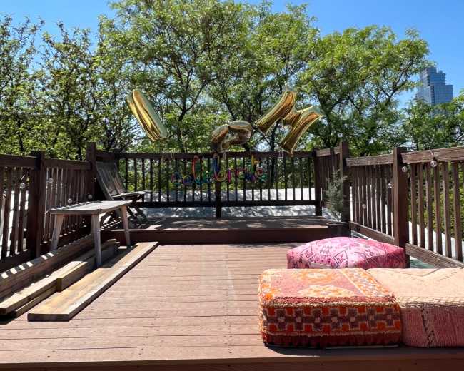 A wooden deck is decorated with colorful pillows and large gold balloons against a clear blue sky and lush green trees.