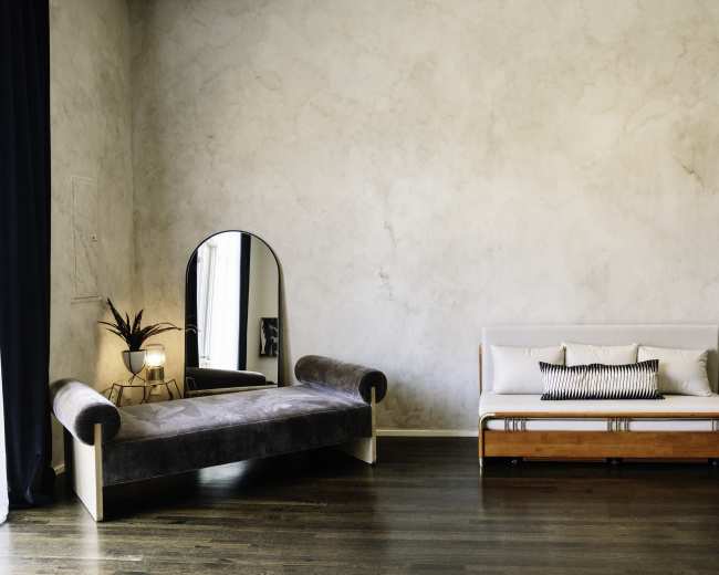 The image shows a minimalist living space featuring a brown chaise lounge, a white sofa with decorative cushions, a round mirror, and a few plants against a textured wall.
