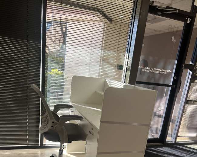 A modern white desk with a chair in a well-lit office space featuring large windows and wooden flooring.