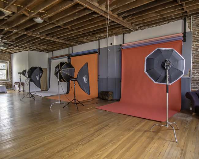 A photography studio with multiple backdrops and lighting equipment set up in a spacious room featuring exposed wooden floors and a high ceiling.