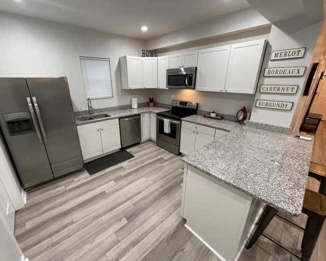 The image shows a modern kitchen with white cabinets, stainless steel appliances, and a granite countertop, featuring a small dining area with wooden stools.