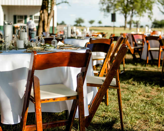 A rustic outdoor dining setup with wooden tables and chairs arranged on grass, beneath a clear sky.