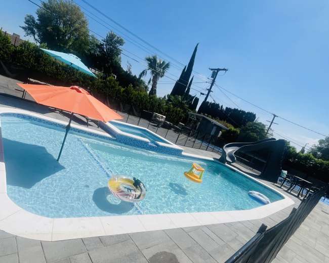 The image shows a swimming pool with a water slide, an orange umbrella, and a floating lounge in a fenced yard.
