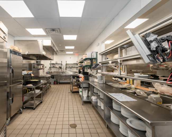 The image shows a well-organized commercial kitchen with stainless steel appliances, workstations, and neatly stacked plates.