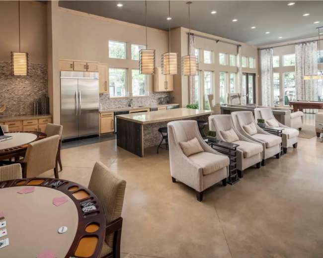 The image shows a spacious common area featuring a card table, a modern kitchenette with stainless steel appliances, and several comfortable chairs arranged in a light-filled room.