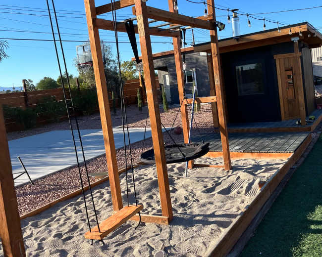 A wooden swing set with a sand base is situated beside a small building in a sunny outdoor area.