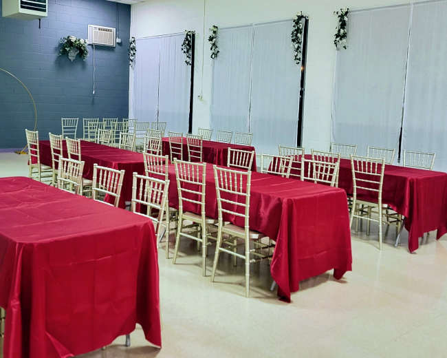 A hall with multiple tables covered in red tablecloths and gold chairs arranged in a grid pattern.