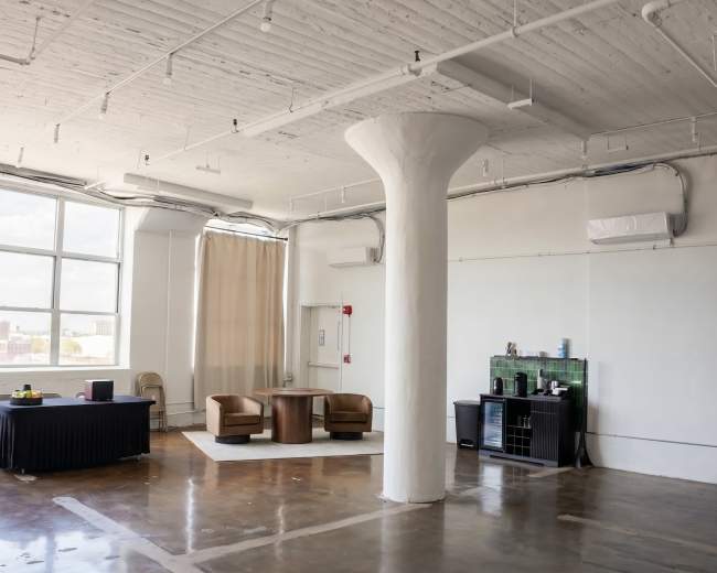 The image shows a spacious, airy room with large windows, featuring a seating area with two chairs and a table, alongside a small kitchenette and a column in the center.