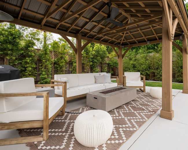A covered outdoor seating area features a sectional sofa, a coffee table, and decorative poufs on a patterned rug.
