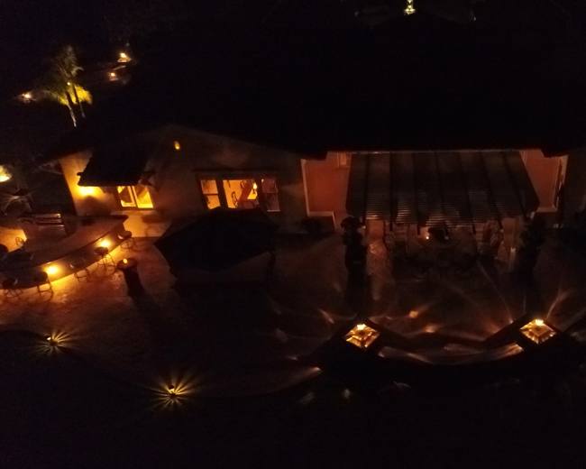 A well-lit outdoor gathering is taking place around a house, surrounded by illuminated pathways and soft lighting in the evening.