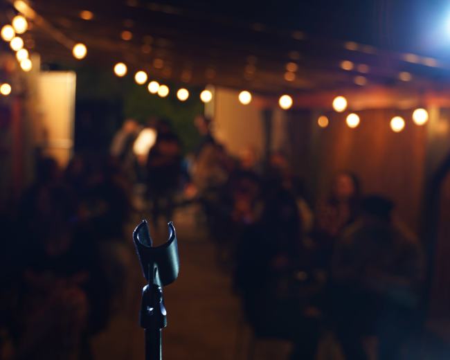 A microphone stands in the foreground, facing an audience seated in a dimly lit space adorned with string lights.