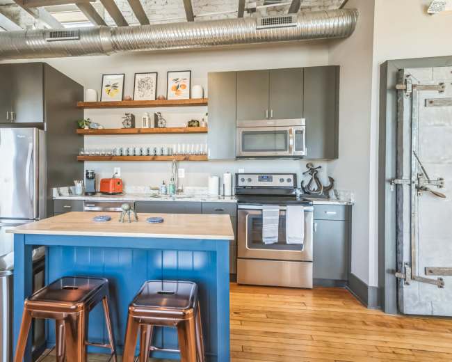 The image shows a modern kitchen featuring stainless steel appliances, dark cabinetry, and a central island with two metal stools.