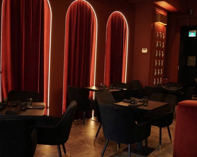 The interior of a restaurant features dark velvet chairs and tables arranged beneath large, arched windows framed with red curtains.