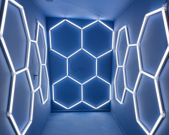 The image shows a narrow hallway with blue walls and illuminated hexagonal neon light fixtures arranged on both sides.