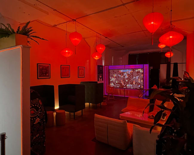 The image depicts a dimly lit lounge area featuring red lanterns, green plants, and a projection screen displaying colorful visuals.