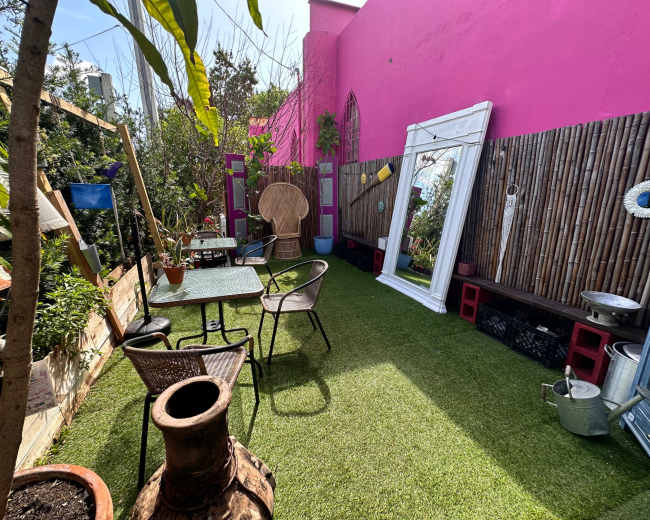 The image shows a vibrant outdoor space with green artificial grass, several small tables and chairs, a large mirror, and a pink wall in the background.