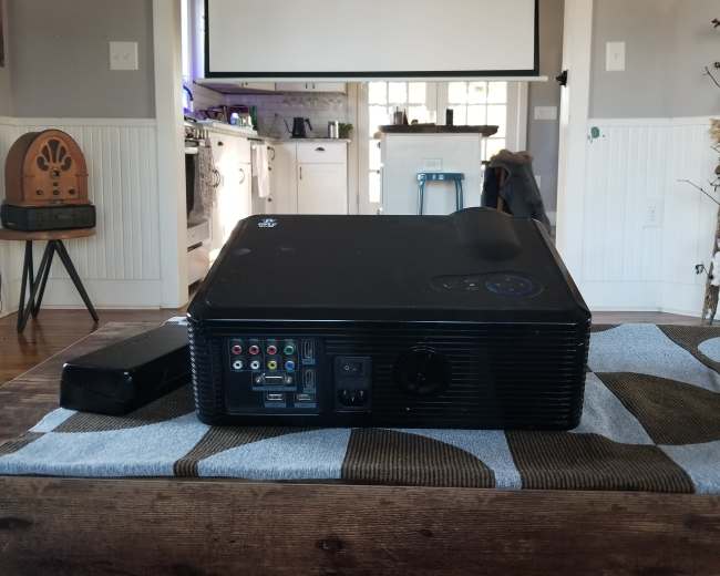 A projector is placed on a wooden table in a room with a large screen in the background.