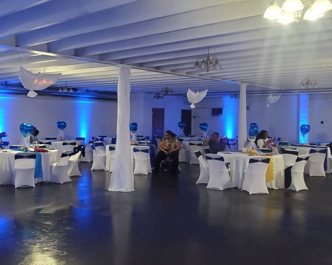 A banquet hall with decorated tables covered in white tablecloths and blue accents, illuminated by overhead lights and blue wall lighting.