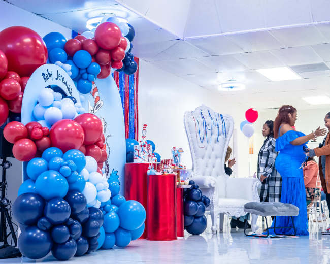A decorated event space features a colorful balloon arch, a cake table, and guests engaged in conversation.