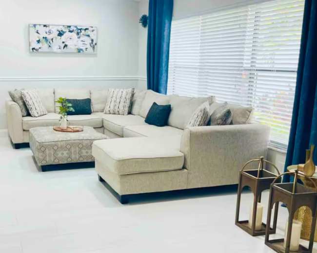 The image shows a neatly arranged living room with a light-colored sectional sofa, a coffee table, and decorative elements including a plant and lanterns.