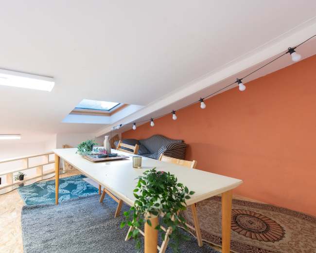 An attic space features a long table surrounded by chairs, with a cozy seating area and decorative plants under a skylight.